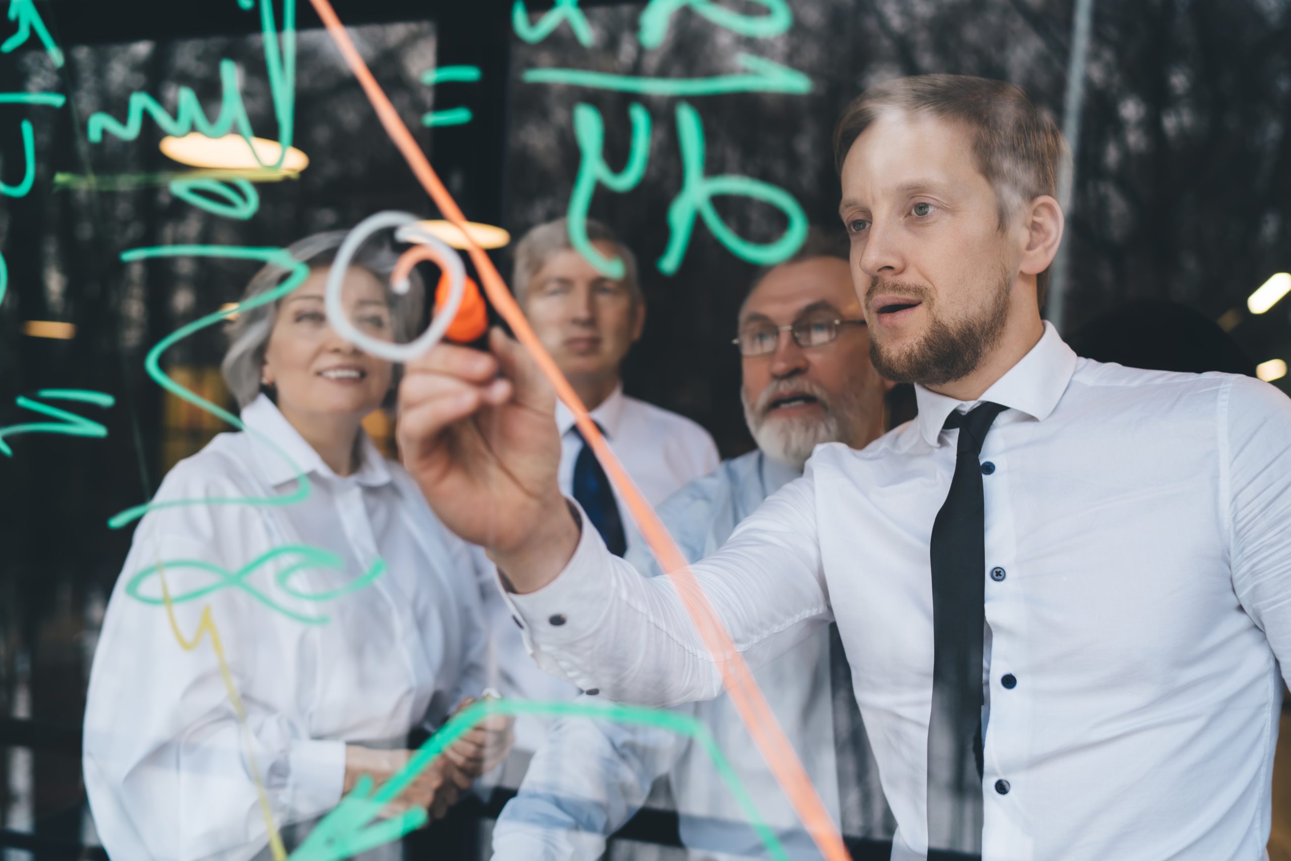 Business partners solving financial strategy together Group of businesspeople in formal outfits solving strategy while young bearded colleague writing on glass of window in conference hall